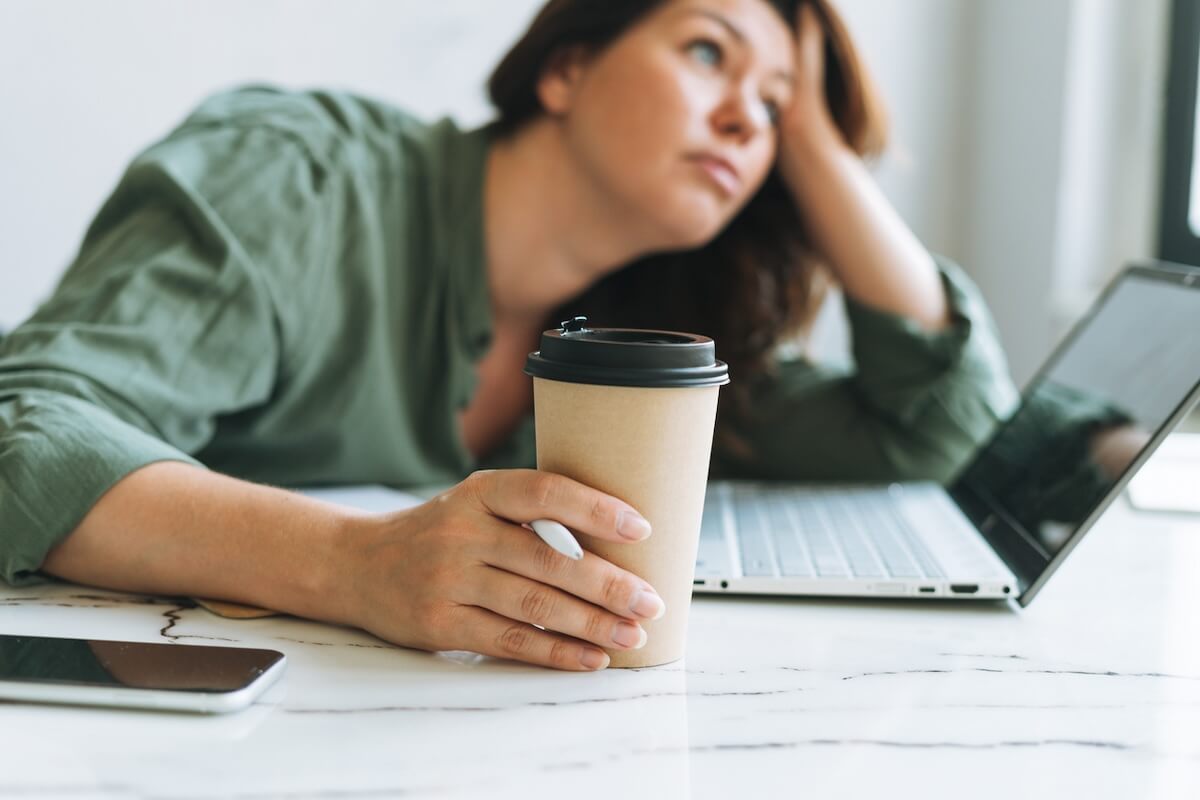 A person in a green shirt leans on a desk, holding a coffee cup in one hand while looking into the distance. A laptop and smartphone are on the desk.