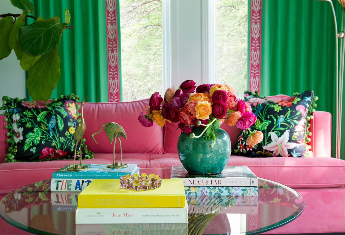 A living room with a pink sofa, floral pillows, green curtains, a glass coffee table adorned with books, a vase of colorful flowers, and various decorative items.